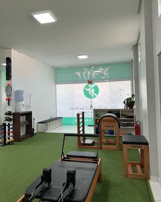 Sala ampla com aparelhos de pilates, grama sintética e o logo da clínica na vitrine.
