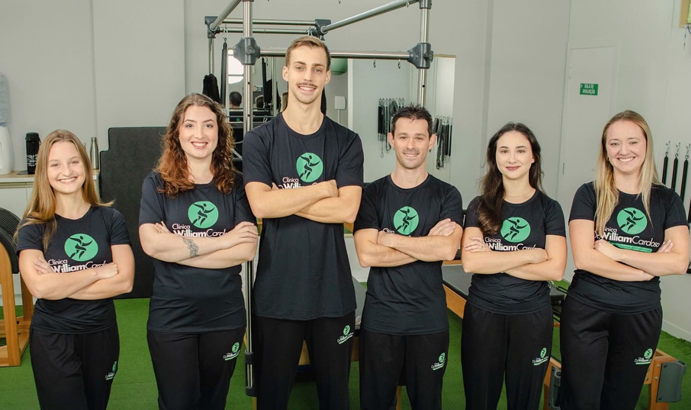 Equipe da clínica, seis fisioterapeutas de camiseta preta com o logo, de braços cruzados na área de treino.